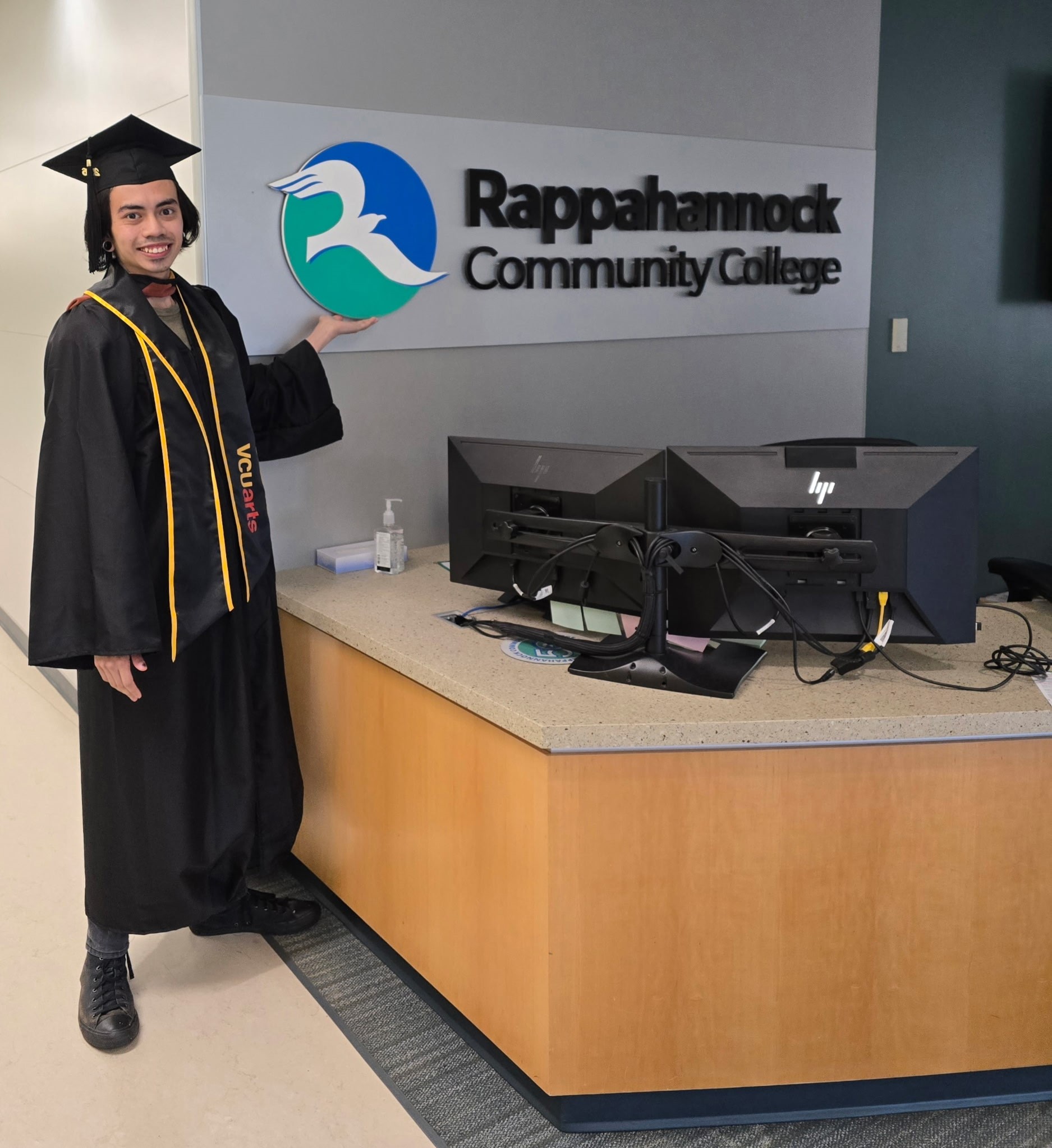 Graduate in cap and gown poses next to the Rappahannock Community College sign in a campus lobby.